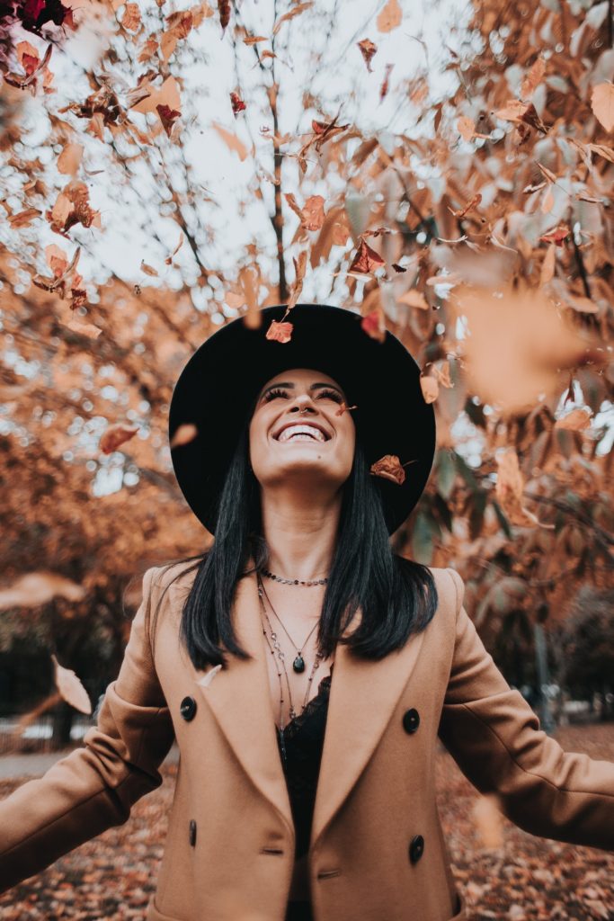 Mulher sorridente de casaco marrom, blusa e chapéu pretos em meio a um parque durante o outono com folhas caindo sobre ela