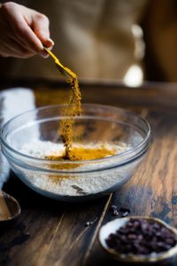 foto horizontal de uma pessoa colocando uma colher de chá de cúrcuma em um bowl de vidro cheio de farinha de trigo