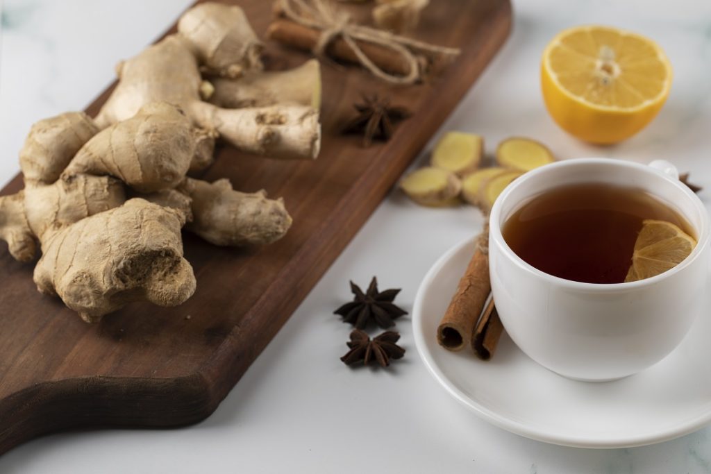 Xícara de chá com limão em cima de uma mesa com toalha branca e, ao lado, uma placa de madeira com pedaços de gengibre e limão