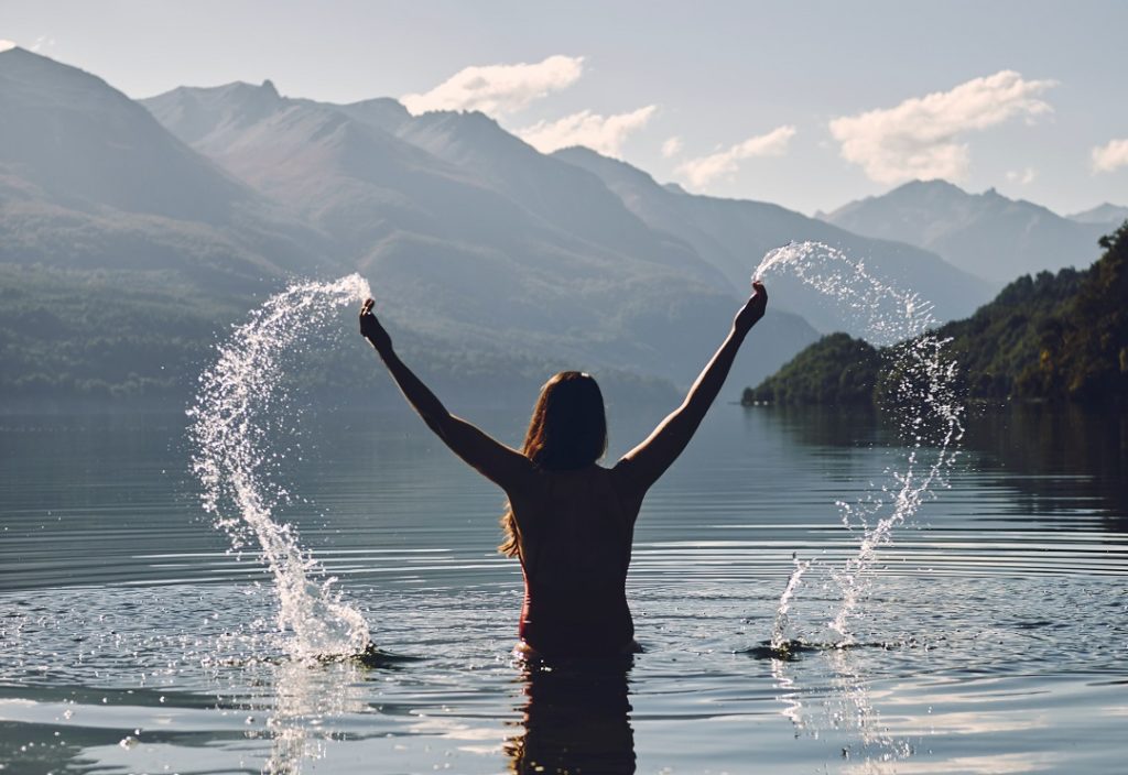 Maha Gunas: mulher em um lago, de costas ao por do sol levntando água coma s mãos em uma paisagem montanhosa